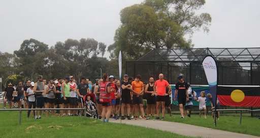 A diverse group of runners gathers at a park, preparing for a race, with an Aboriginal flag displayed in the background.