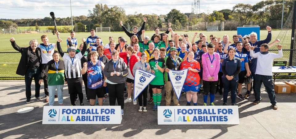 A diverse group of athletes and supporters pose happily with banners for "Football for Abilities" at a sports field event.