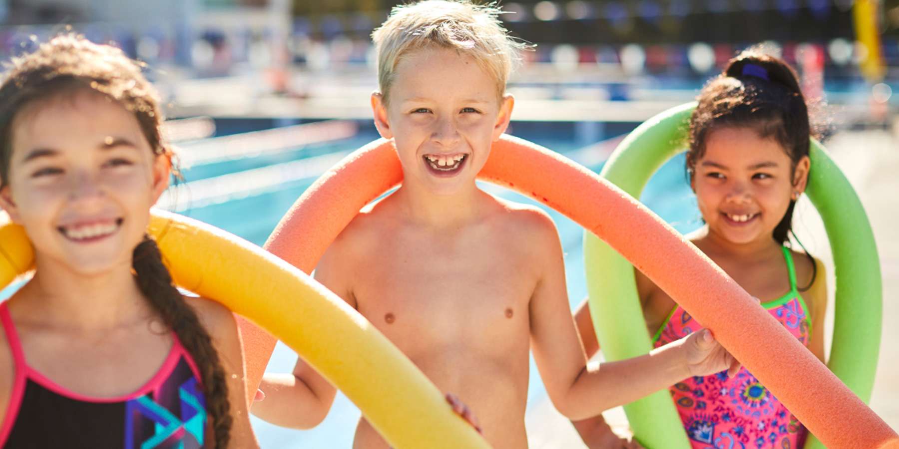Three children, two girls and a boy, stand together with colorful pool noodles at a sunny swimming pool.