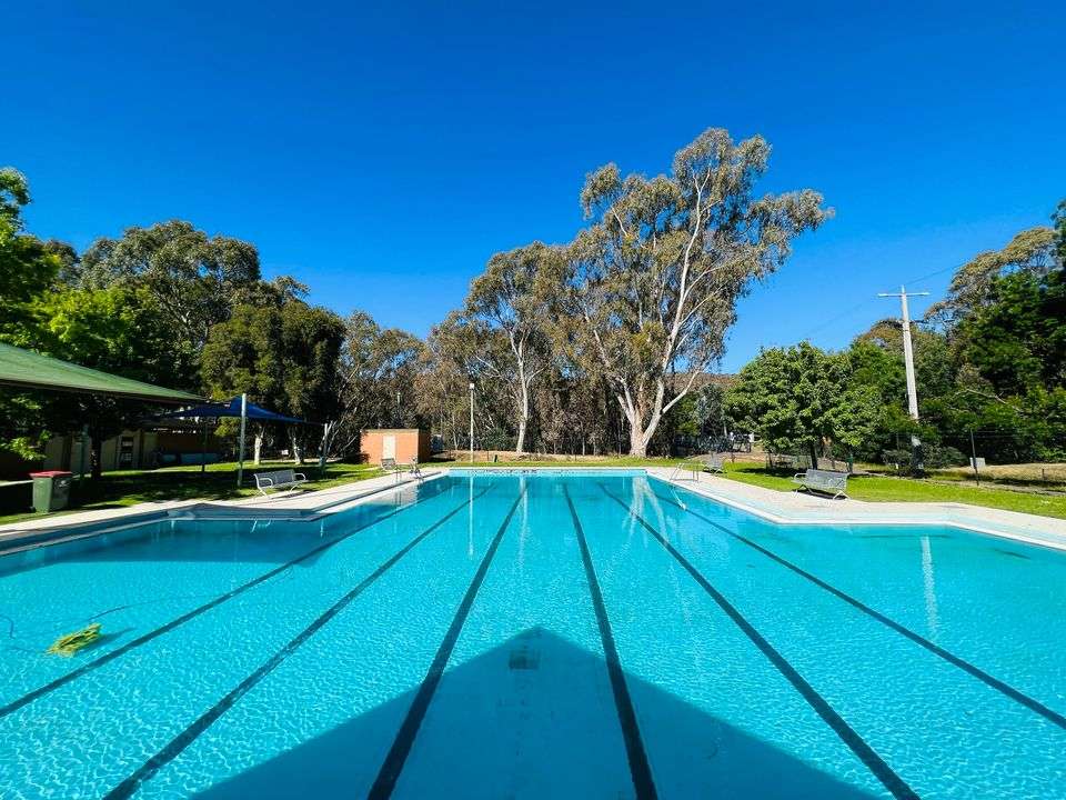 large inground swimming pool, with beige shade sail surrounded by green grass and trees.