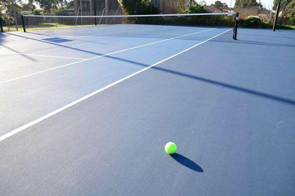 A large tennis court with net with a blue surface and 1 x green tennis ball on the ground.