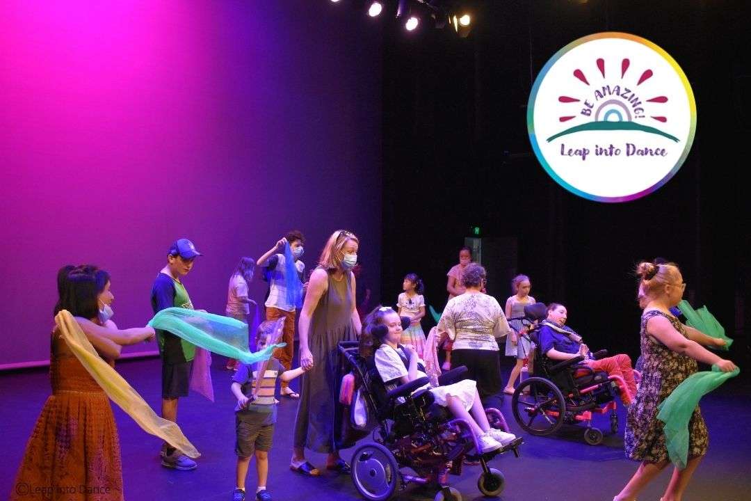 A vibrant dance performance with participants of all ages, including those in wheelchairs, engaging with colorful scarves under purple lights.