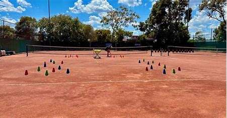 Image displays a large tennis court with a red sand court, you can see a blue sky with clouds, and trees in the background