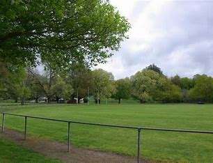Image displays a green oval with a path, a fence and trees in the background. The sky is grey and overcast in this image