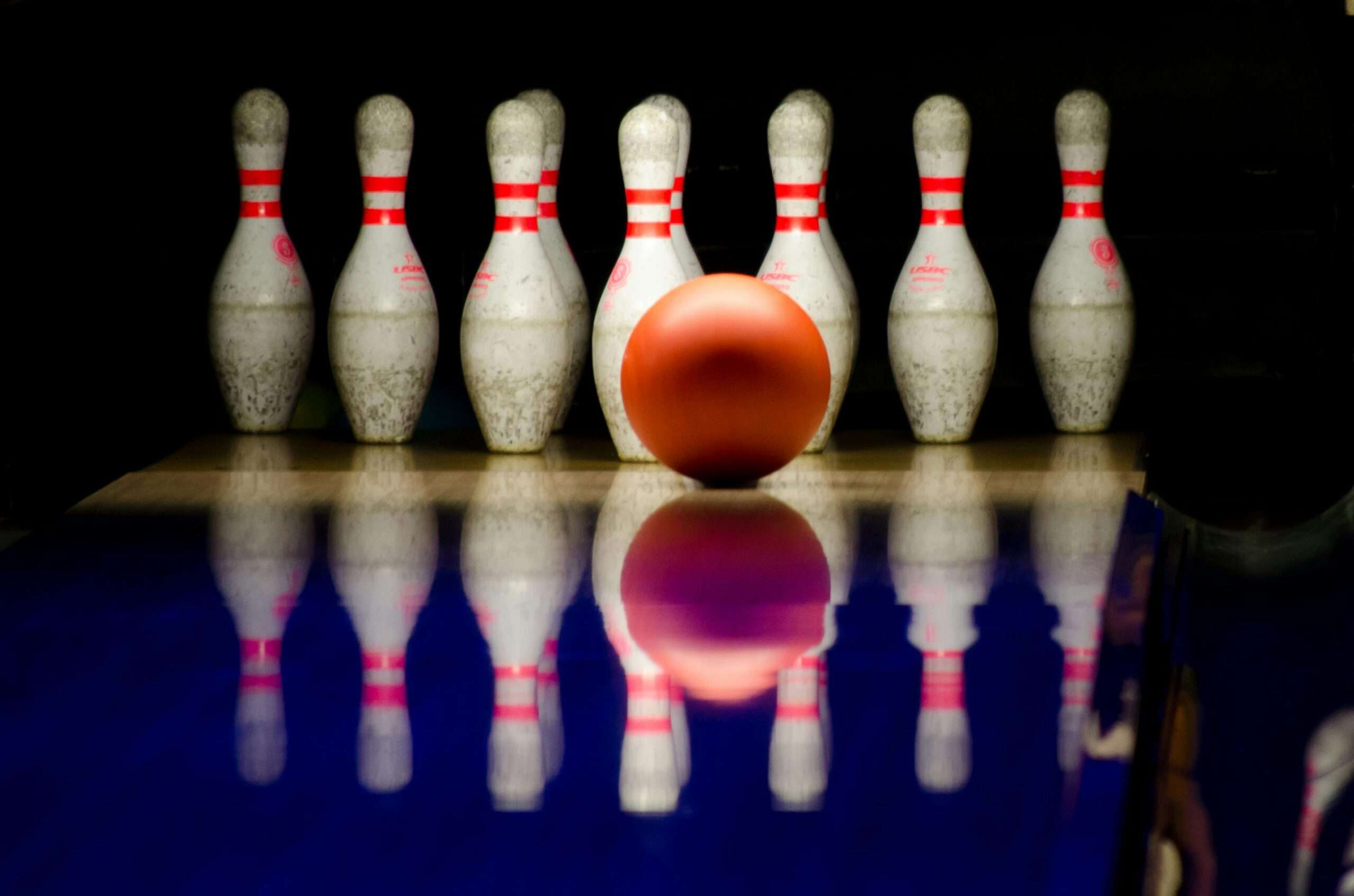 An orange bowling ball rolling toward a set of white pins with red stripes, reflected on a smooth blue lane.
