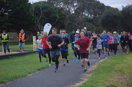 A large group of runners in athletic gear takes off at the start of a park run, surrounded by trees and green grass.