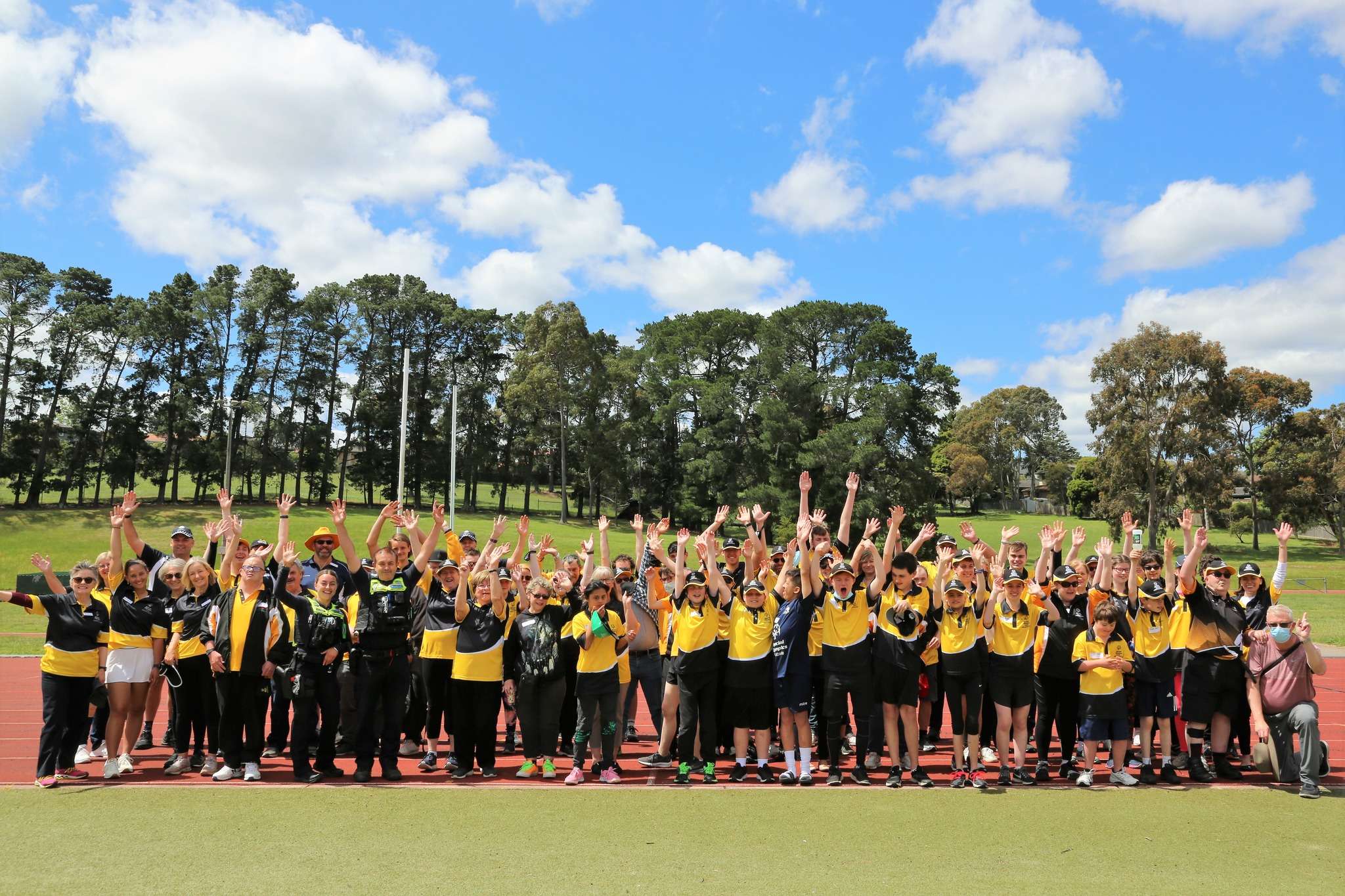 A large group of cheery individuals in yellow shirts stands outdoors, with hands raised in celebration against a sunny blue sky.