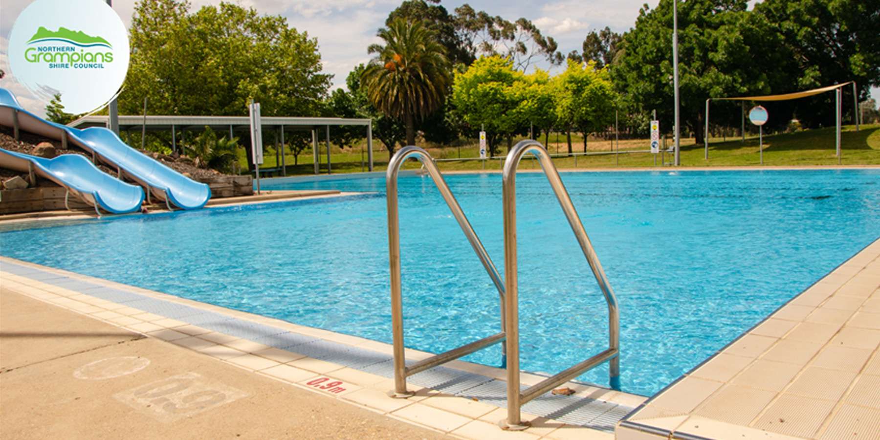 Blue swimming pool with a metal ladder and water slides, surrounded by lush greenery and clear skies.