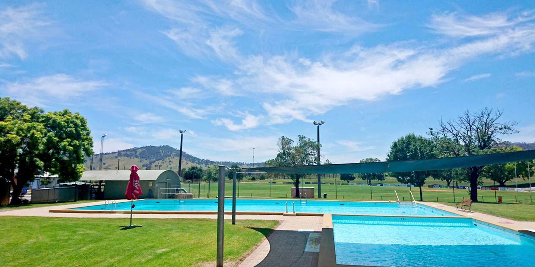 A bright, sunny day at a swimming pool surrounded by greenery and mountain views, featuring blue water and a red umbrella.