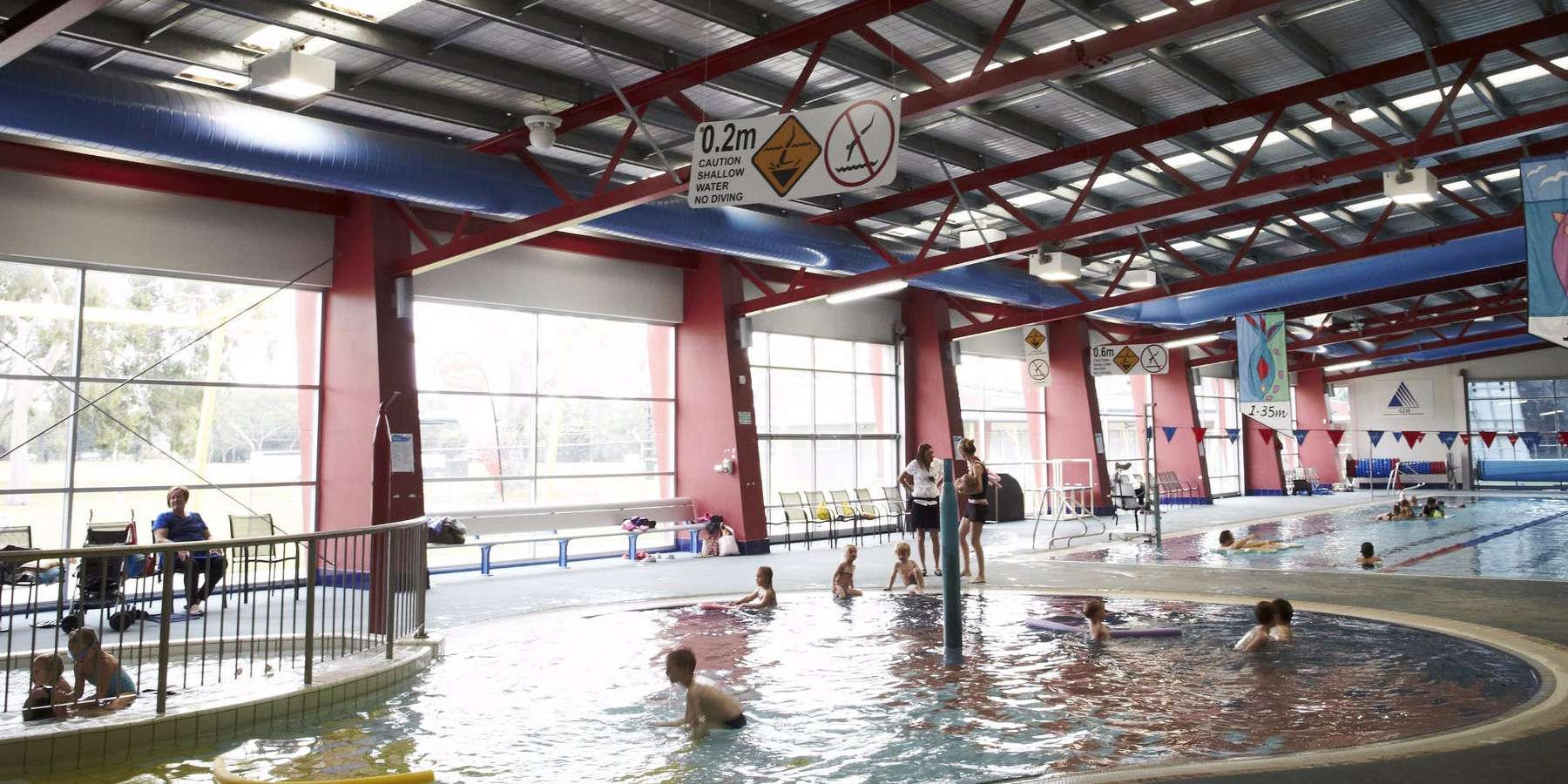 An indoor swimming pool with children and adults enjoying the water under bright, spacious windows and a colorful ceiling.