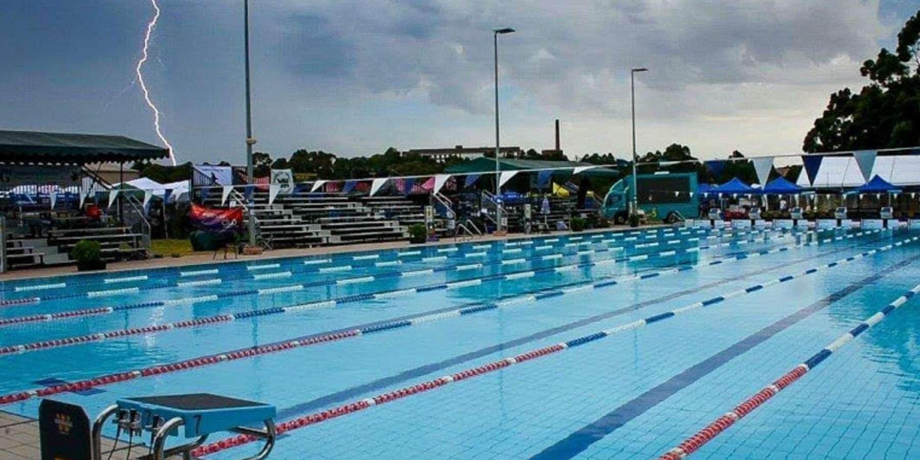 A stormy sky looms over an empty swimming pool, with lightning visible in the background and spectators under tents nearby.