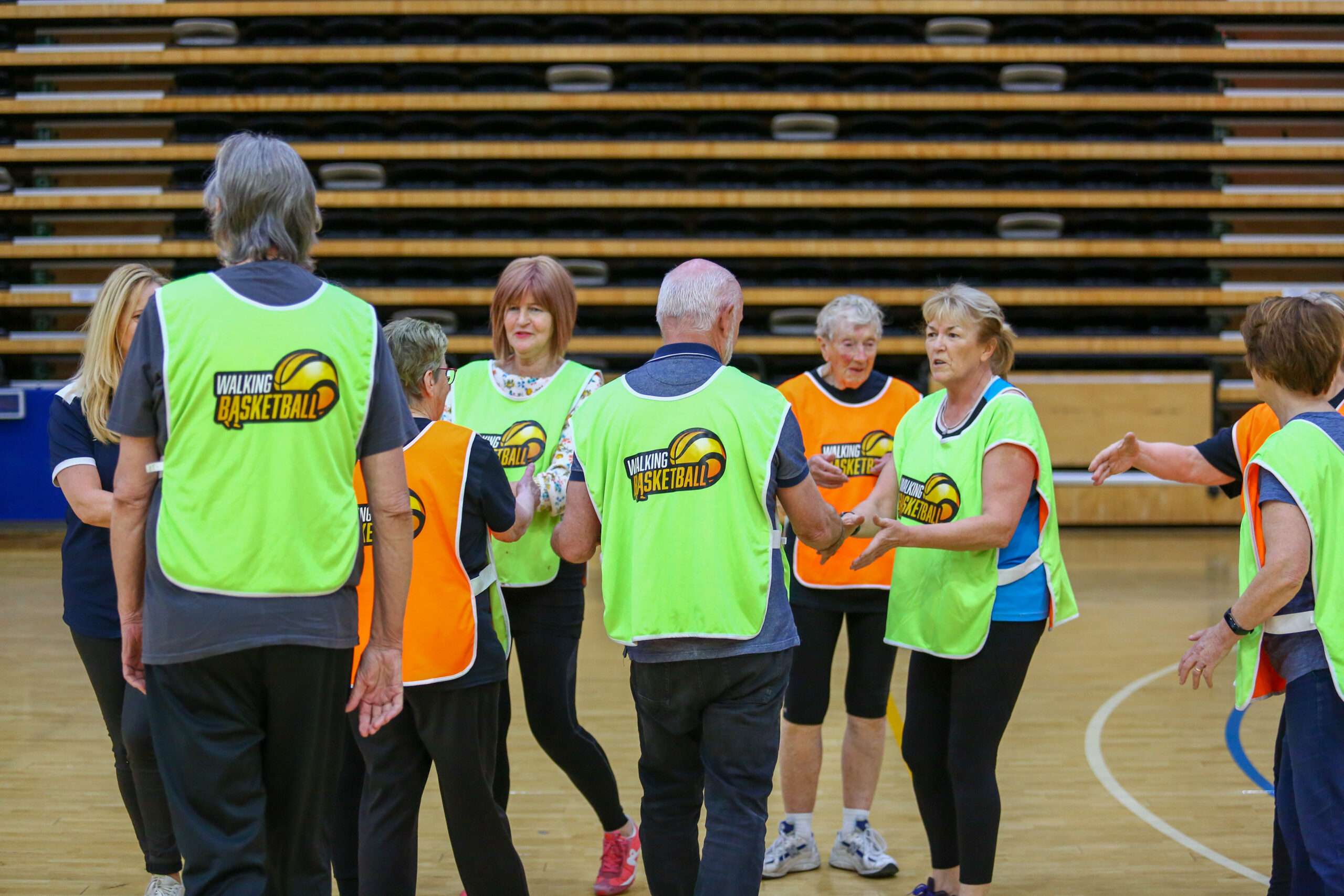 A group of older adults in neon jerseys engage in a sports activity in a gymnasium, smiling and interacting with each other.