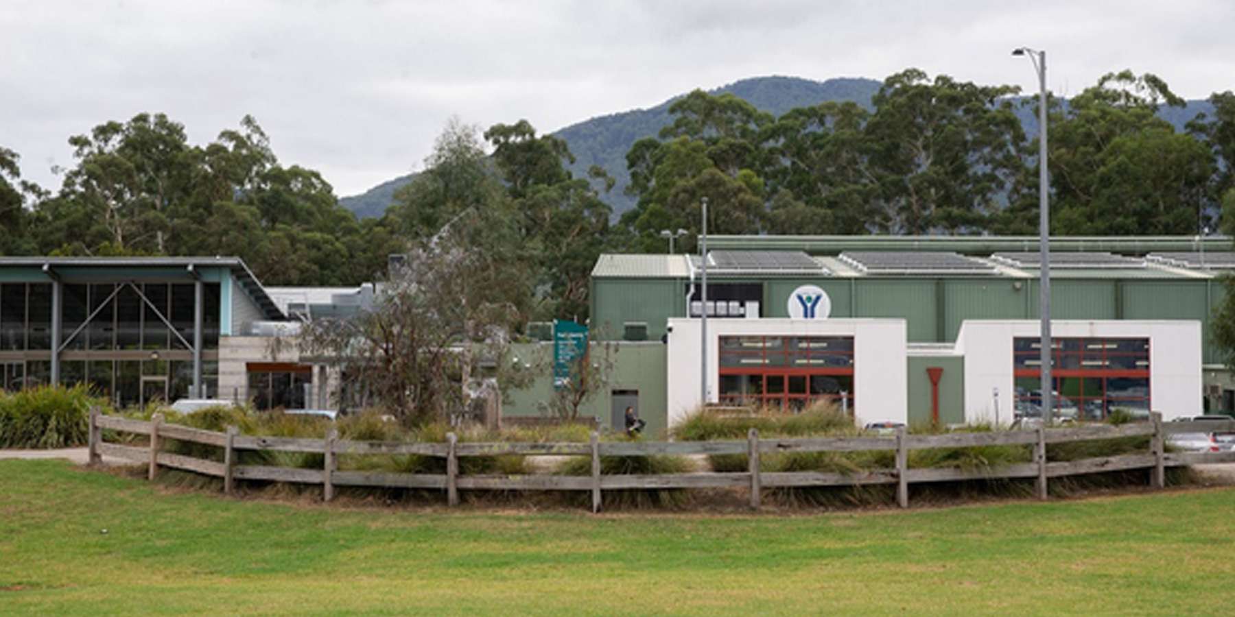 Modern building with a green exterior, surrounded by landscaped gardens and mountains in the background under a cloudy sky.